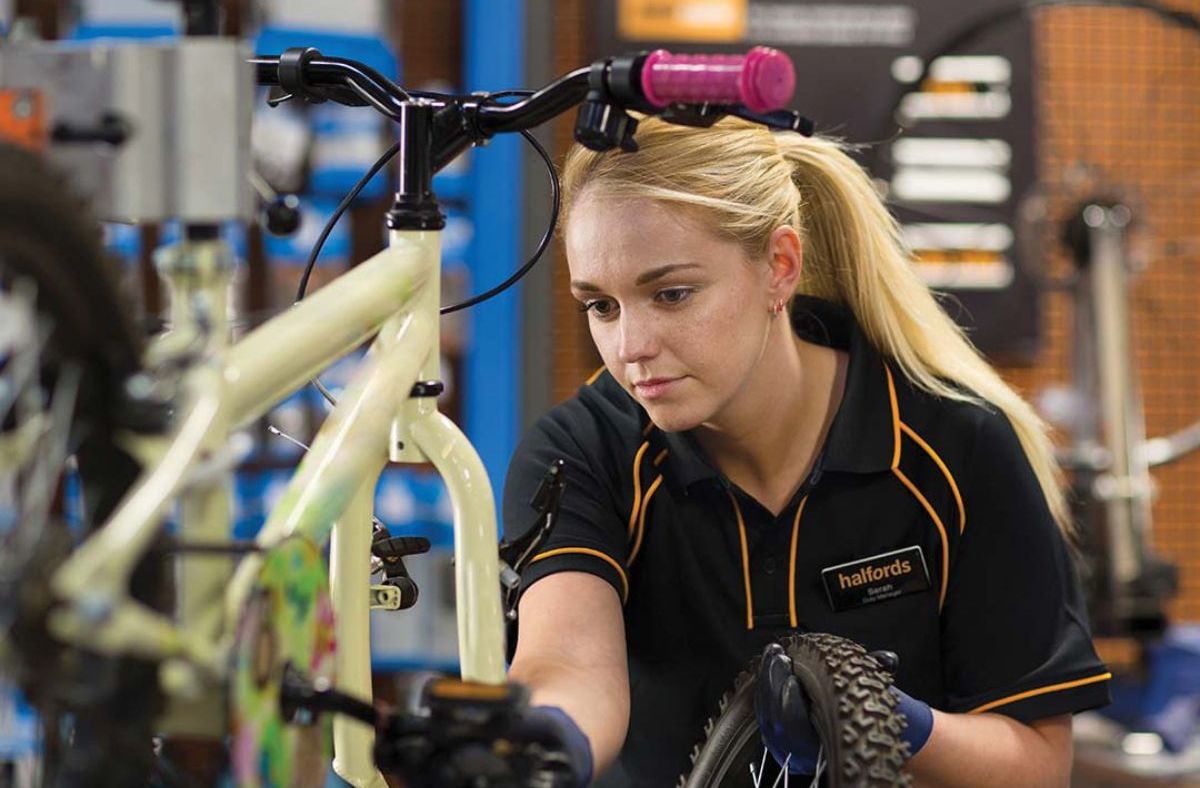 Halfords technician Sarah in a black polo shirt with orange trim, assembling a bicycle in a workshop.