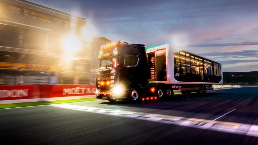 Black Formula 1 hospitality truck driving on a race track at dusk, bright lights and sponsor banners visible.
