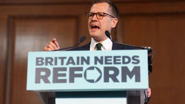 Robert Jenrick speaking at a podium with BRITAIN NEEDS REFORM sign, delivering a speech.