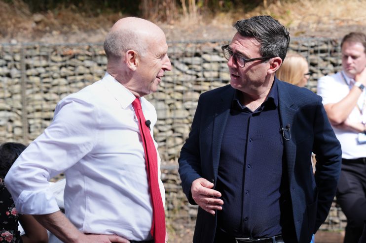 John Healey, in a red tie, speaking with Andy Burnham, wearing glasses and a dark blue jacket, outdoors.