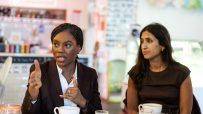 Kemi Badenoch speaks, gesturing with hands, while Claire Coutinho listens intently at a table with coffee cups.