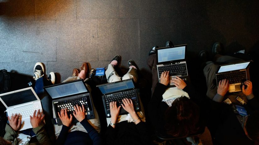 Overhead view of several laptop workers typing on computers, seated on a dark floor.