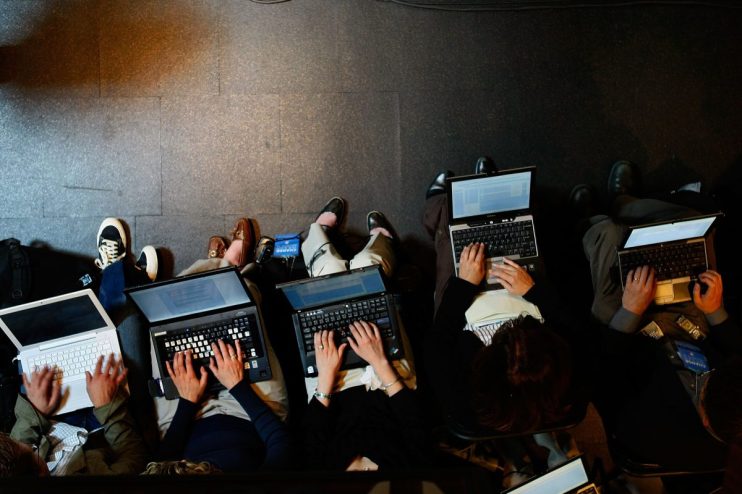 Overhead view of several laptop workers typing on computers, seated on a dark floor.