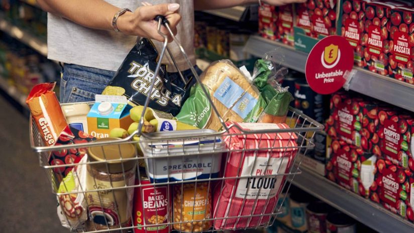 Shopper holding a basket filled with Morrisons groceries, including tortilla chips, bread, and flour, next to an Unbeatabl...