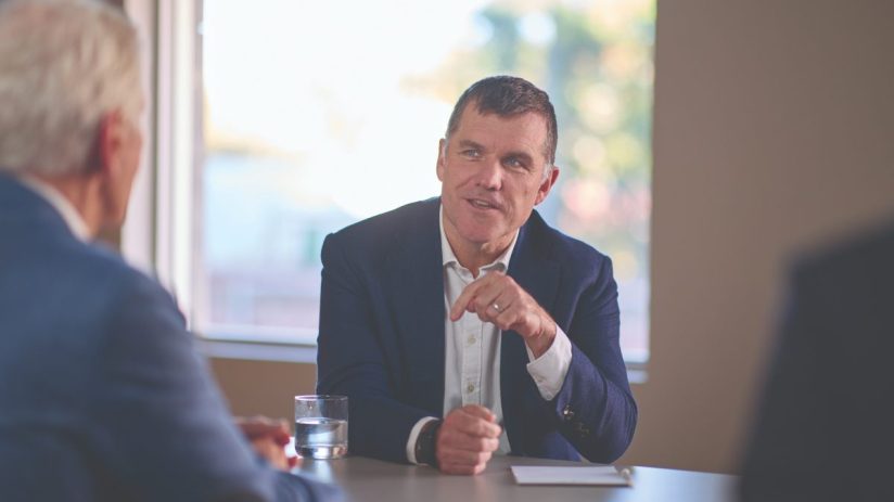 Paul Cooper, a man in a navy suit, gestures during a business meeting at a conference table.