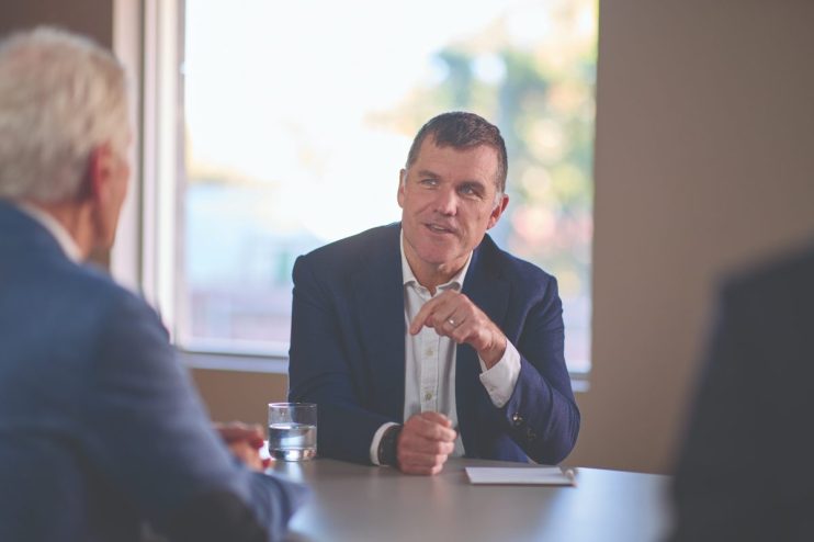 Paul Cooper, a man in a navy suit, gestures during a business meeting at a conference table.
