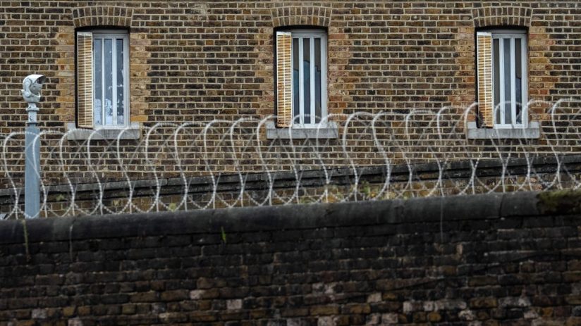 Brick prison wall with barred windows, razor wire, and a security camera.