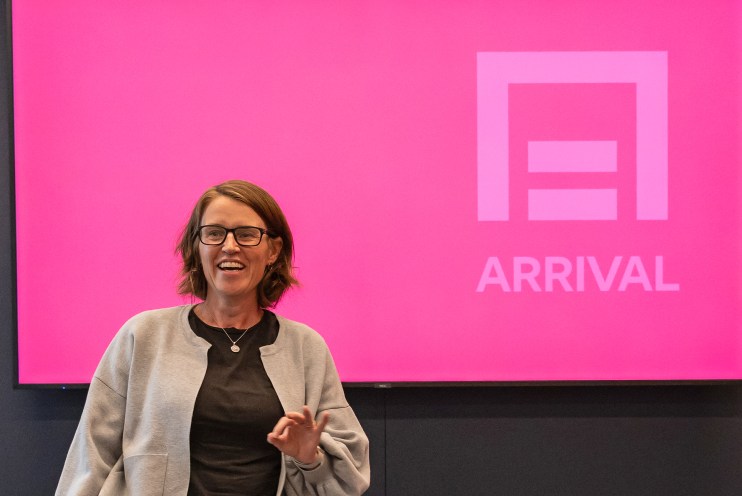 Smiling woman in glasses giving a presentation in front of a bright pink ARRIVAL screen.