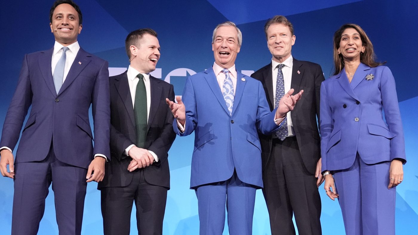 Reform UK leaders Nigel Farage, Richard Tice, and others smiling on stage against a blue background