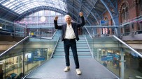 Richard Branson with arms raised in victory on a modern staircase inside St. Pancras International Station