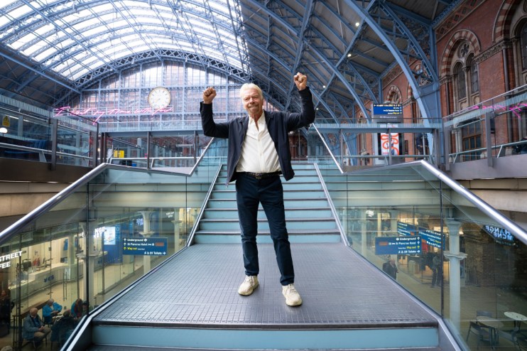 Richard Branson with arms raised in victory on a modern staircase inside St. Pancras International Station