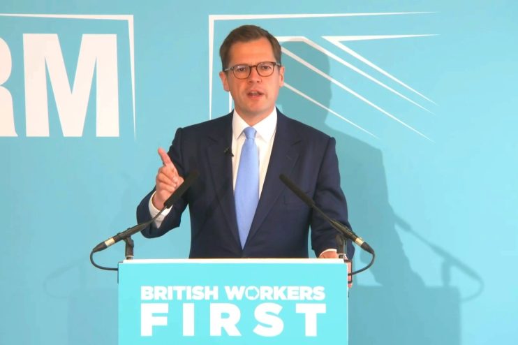 Robert Jenrick speaking at a podium with BRITISH WORKERS FIRST displayed, gesturing with his right hand.