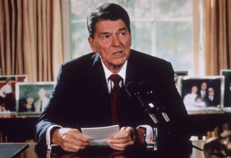 Ronald Reagan speaking at a podium with microphones, holding a paper, family photos visible in the background