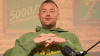 Man in a green hoodie speaking into a microphone, with a 5000 sign in the background.