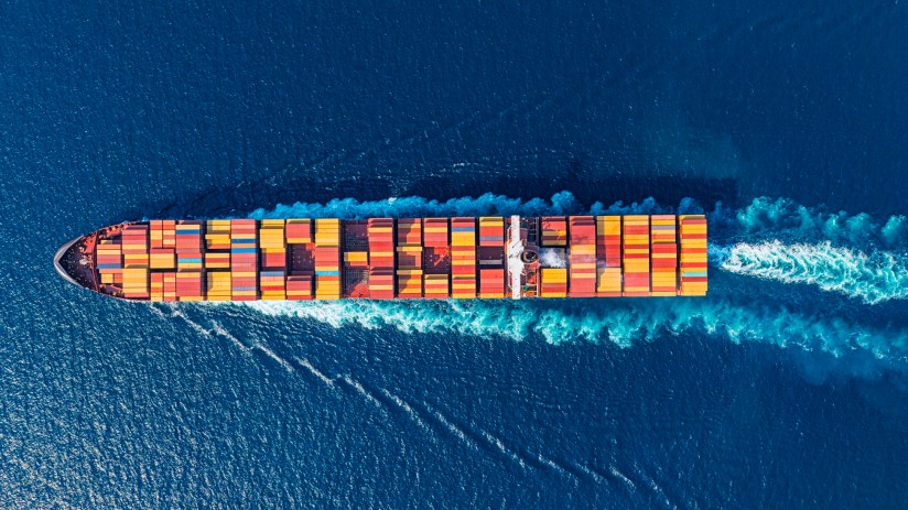 Aerial view of a large container ship moving through deep blue ocean waters, leaving a white wake.