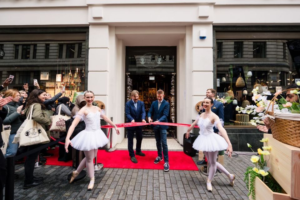 Sostrene Grene store opening with two men cutting a red ribbon, flanked by ballerinas and onlookers on a red carpet.