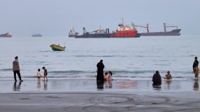 People on a beach with cargo ships and a small boat in the Strait of Hormuz