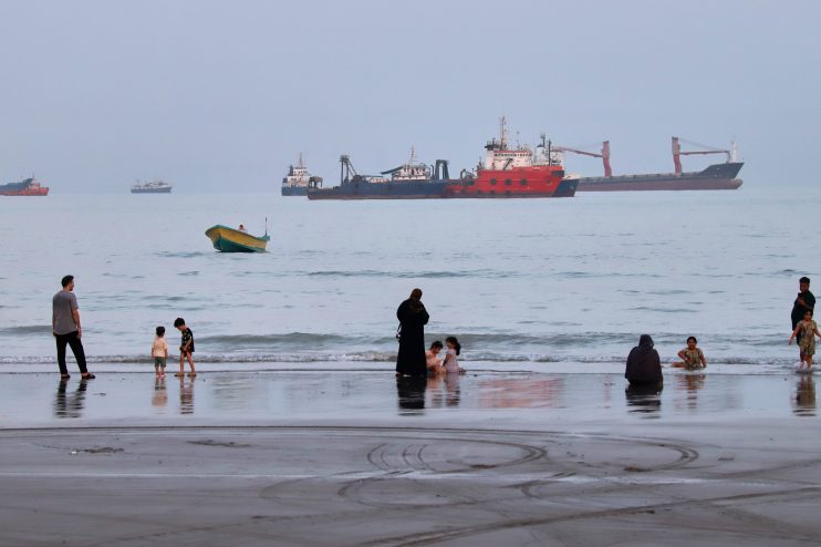 People on a beach with cargo ships and a small boat in the Strait of Hormuz