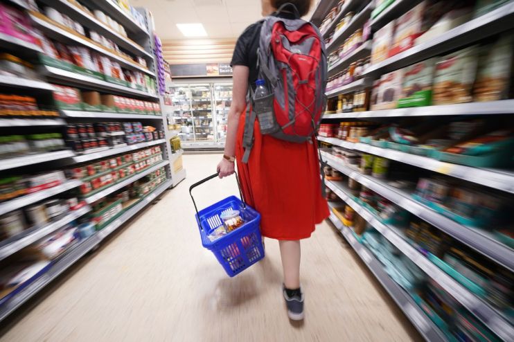 Shopper with red backpack and blue basket walking through a supermarket aisle filled with groceries
