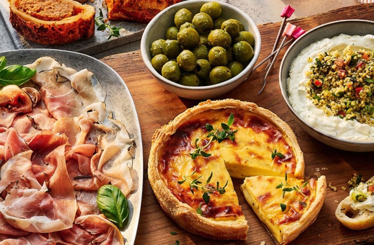Tesco quiche, cured meats, olives, and dip on a wooden board, ready for a party or meal.