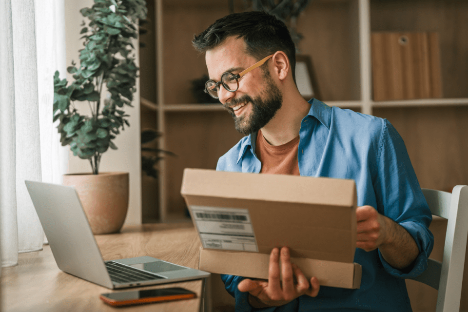 Smiling man with glasses opening a delivery box at a desk with a laptop, reflecting successful online shopping