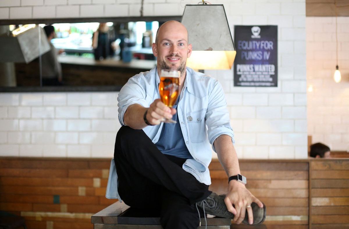 Smiling man in light blue shirt holding up a glass of amber beer in a bar setting