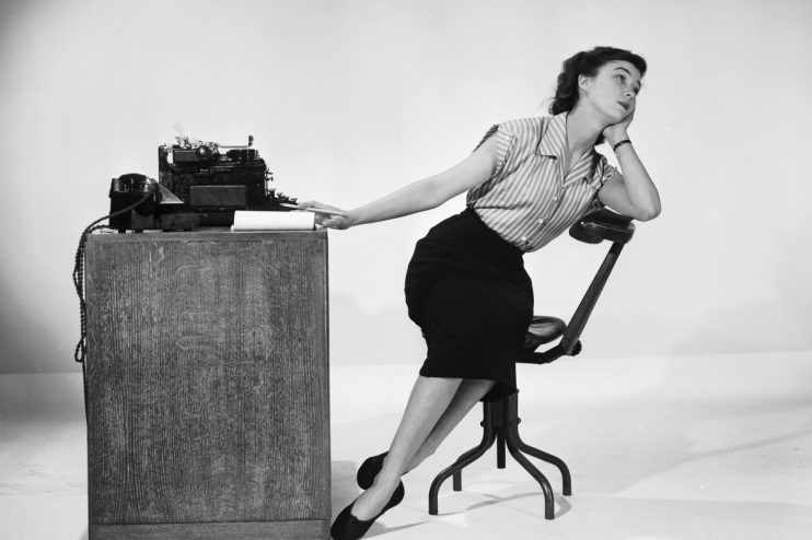 Woman leaning on a desk with a typewriter, looking up thoughtfully, a pencil in hand.