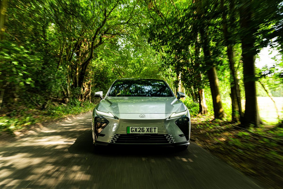 Silver Lexus RZ electric SUV driving on a shaded road through a lush green forest, front view