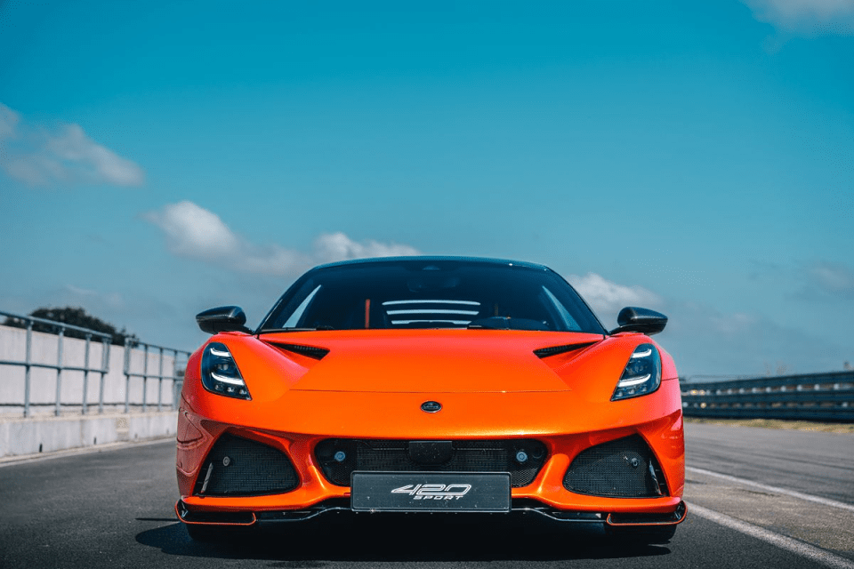 Front view of a bright orange Lotus Emira 420 Sport on a racetrack under a blue sky.