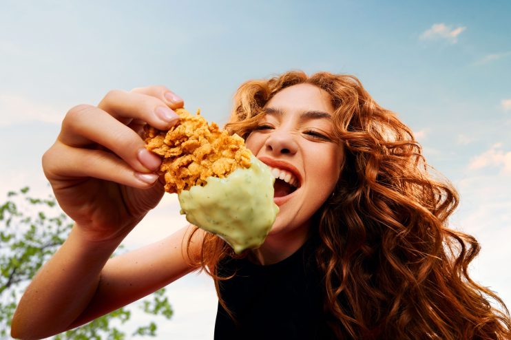 Redhead woman excitedly biting into a piece of KFC fried chicken dipped in green sauce.