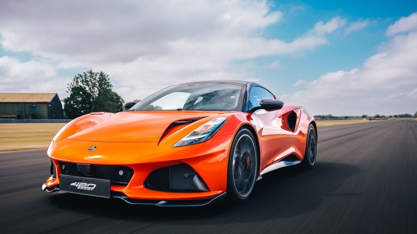 Bright orange Lotus Emira 400 Sport speeding on a racetrack under a blue sky with clouds.