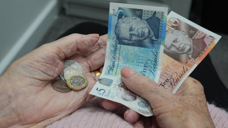Elderly hands holding British pound notes (£5, £10) and coins, representing pension funds and finances.