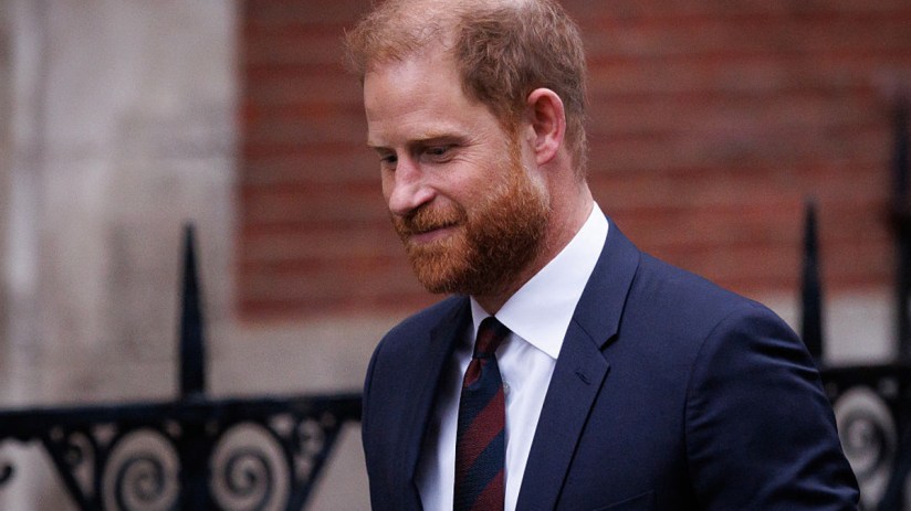 Prince Harry, Duke of Sussex, in a navy suit, white shirt, and striped tie, looking down.