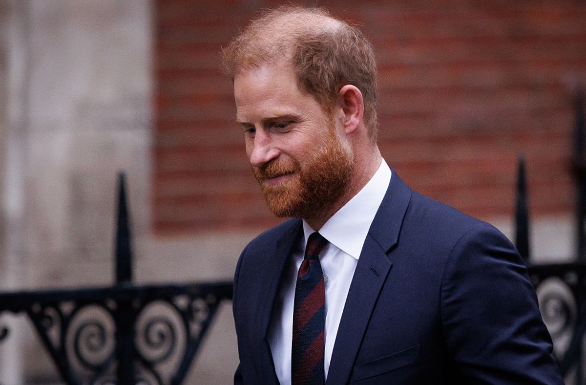 Prince Harry, Duke of Sussex, in a navy suit, white shirt, and striped tie, looking down.