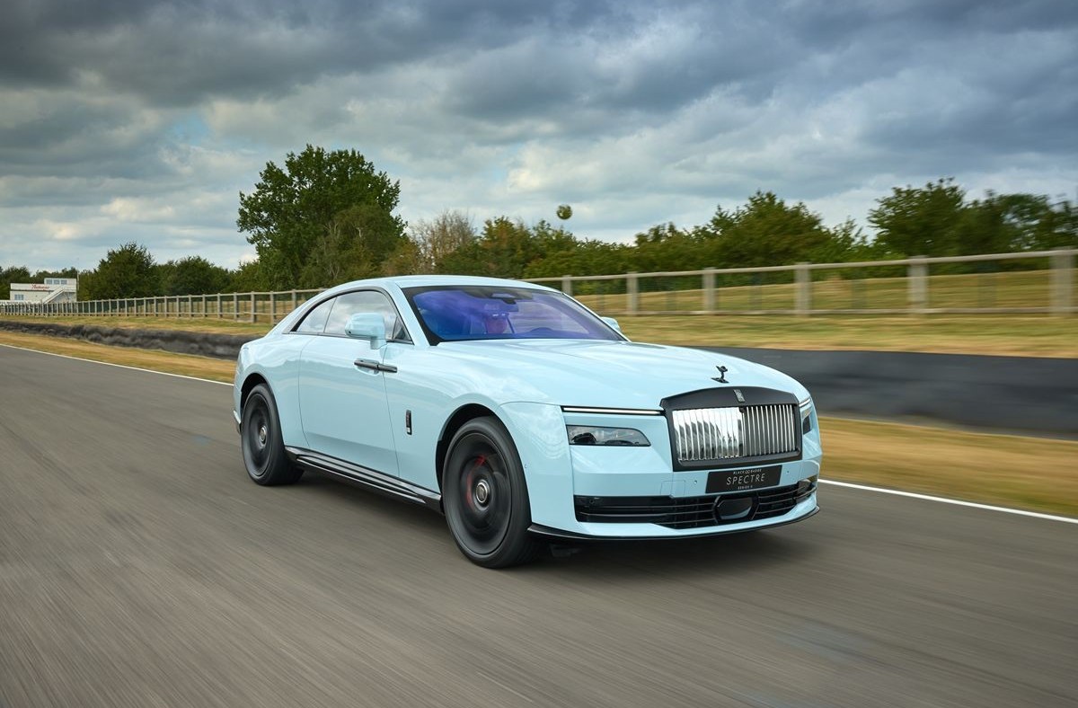 Light blue Rolls-Royce Spectre electric coupe driving fast on a race track under a cloudy sky.
