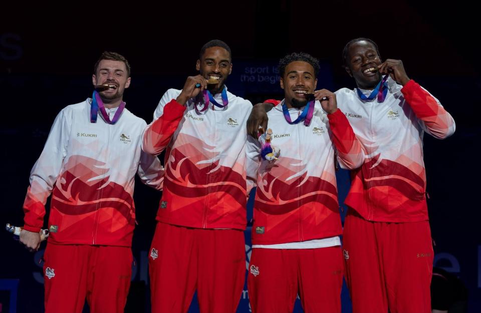 Team England Commonwealth Games gold medalists biting their medals, wearing white and red jackets.