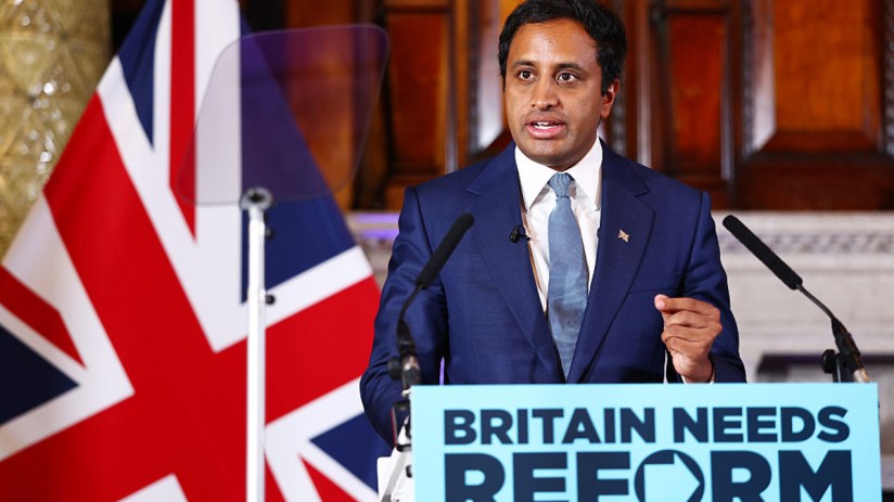 Zia Yusuf speaking at a Reform UK press conference, with a Union Jack flag and Britain Needs Reform sign.