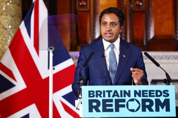 Zia Yusuf speaking at a Reform UK press conference, with a Union Jack flag and Britain Needs Reform sign.