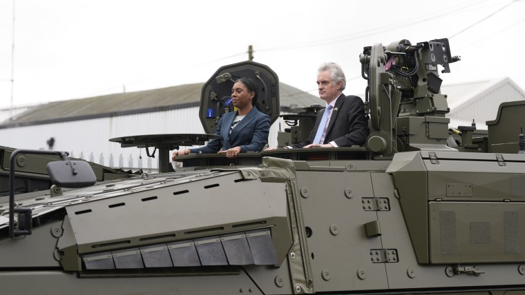 Two people, a woman and a man, standing in the open hatches of a large olive-green military vehicle.