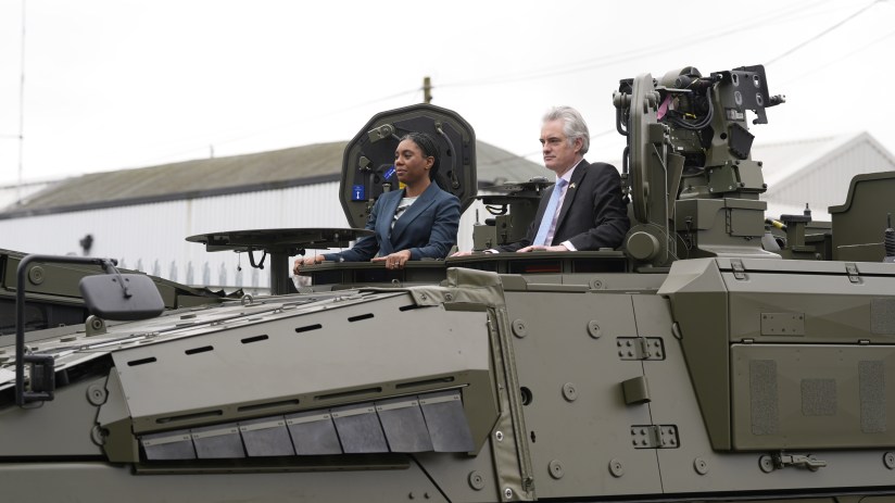 Two people, a woman and a man, standing in the open hatches of a large olive-green military vehicle.