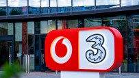 Vodafone and Three company logos on a red and white sign outside a modern glass building