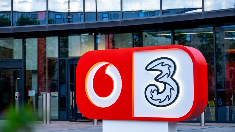 Vodafone and Three company logos on a red and white sign outside a modern glass building