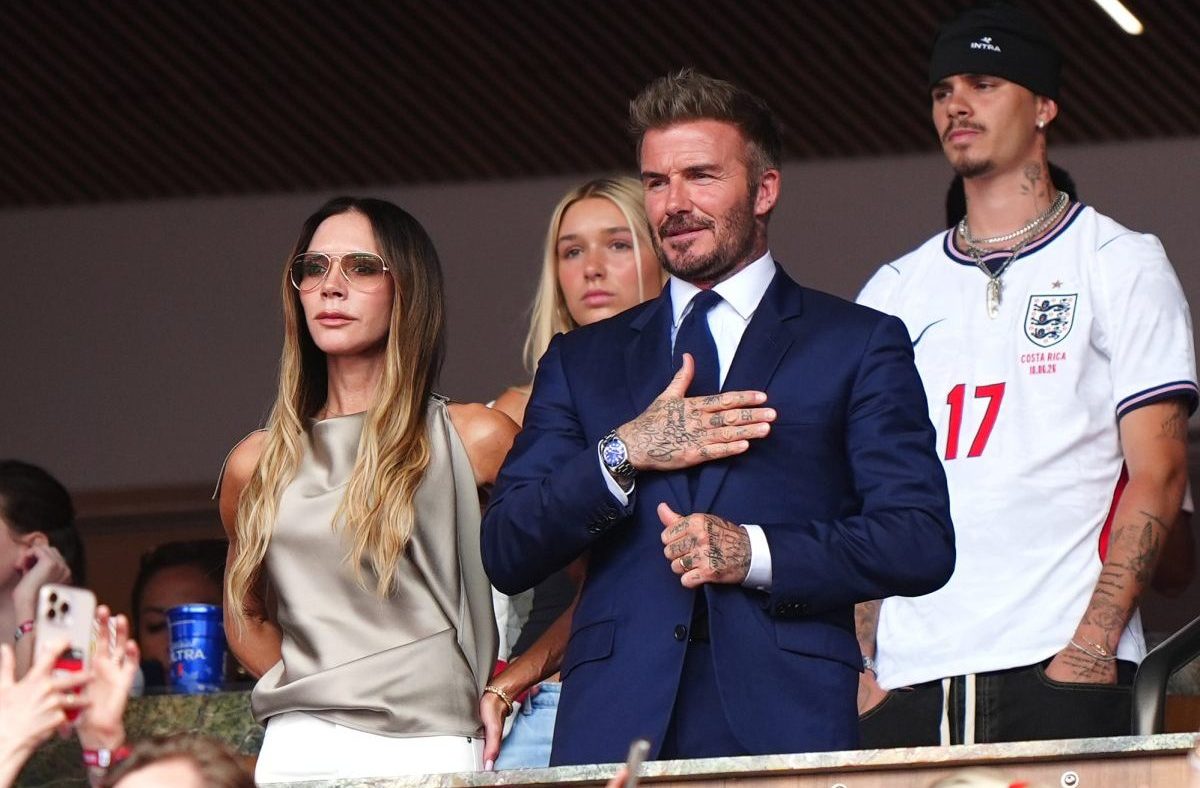 David Beckham in a navy suit with hand on chest, Victoria Beckham in sunglasses, and Brooklyn Beckham in an England jersey.