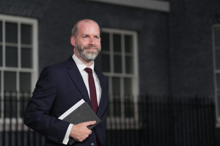 A man with a beard and dark suit holding a notebook, standing in front of a building with windows.