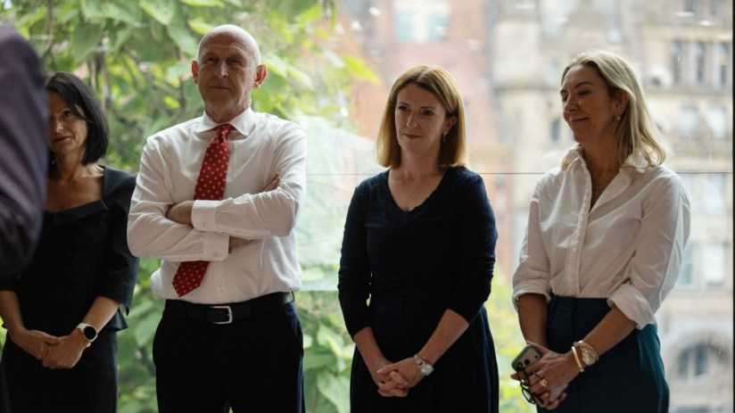 Four professionals, two women and a man, standing indoors with a cityscape visible through a large window.