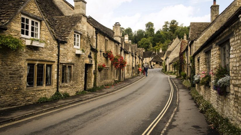 Picturesque English village street with stone cottages and colorful flower boxes, leading to a bend in the road.