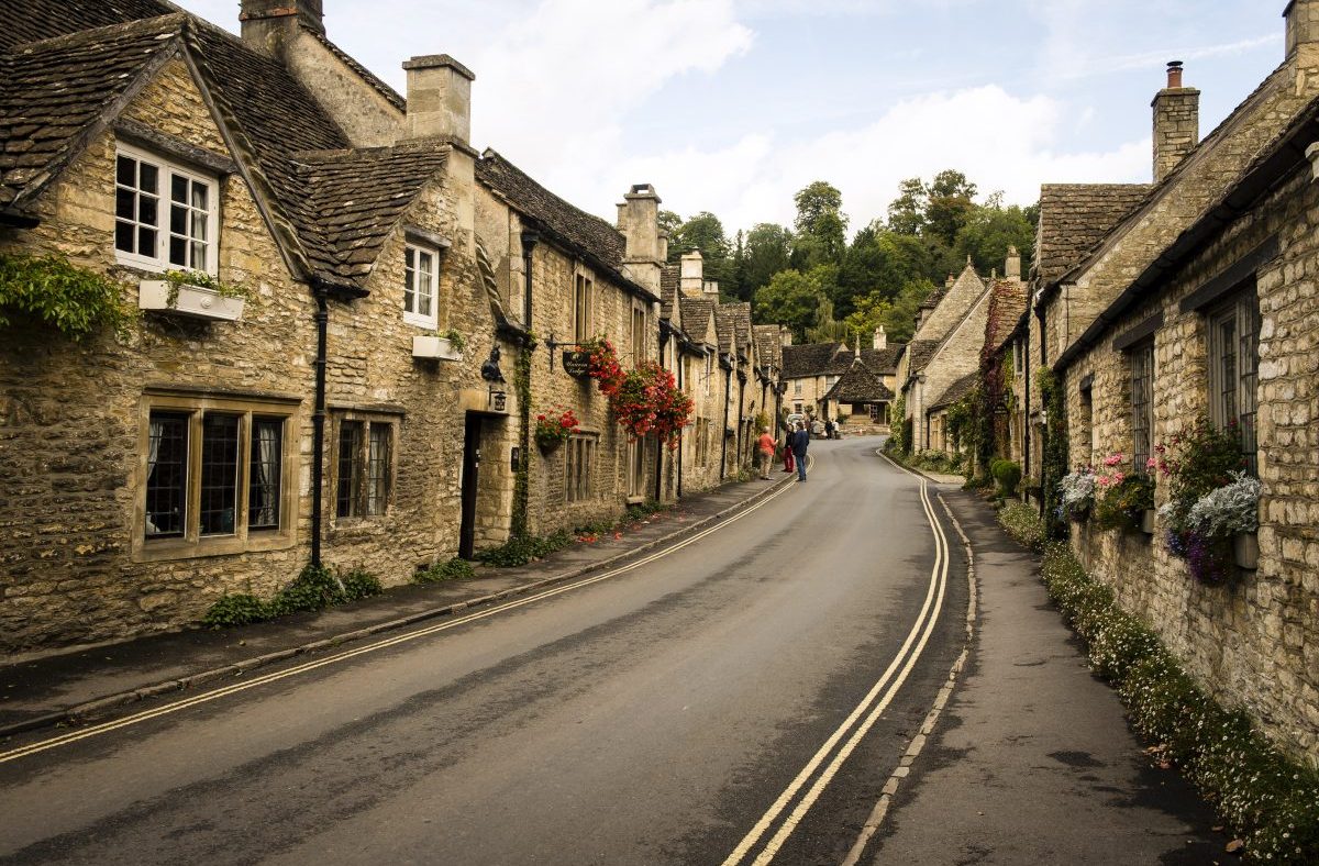 Picturesque English village street with stone cottages and colorful flower boxes, leading to a bend in the road.