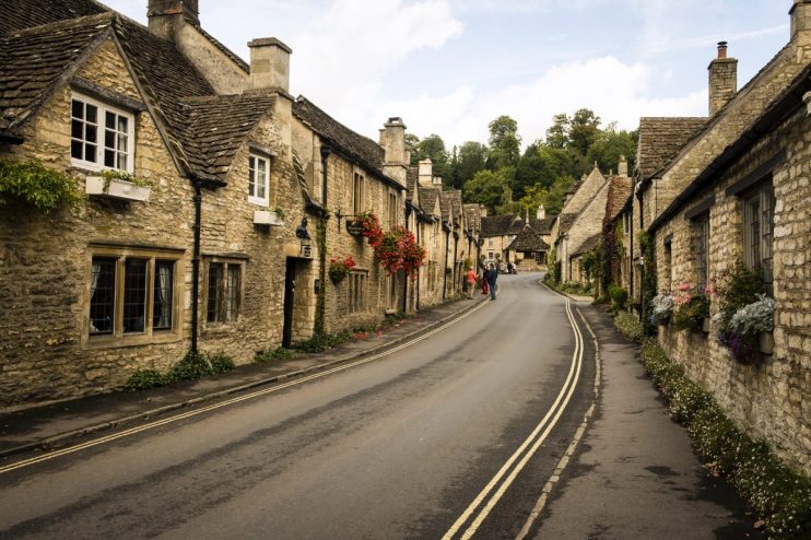 Picturesque English village street with stone cottages and colorful flower boxes, leading to a bend in the road.