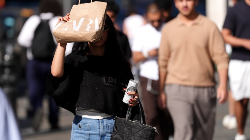 Woman in sunglasses holding a Zara shopping bag over her face, walking in a busy street.