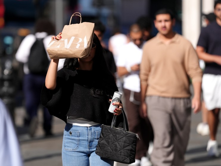 Woman in sunglasses holding a Zara shopping bag over her face, walking in a busy street.